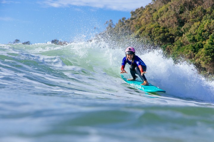 Child in helmet surfing on a wave near a forested shoreline under a blue sky.