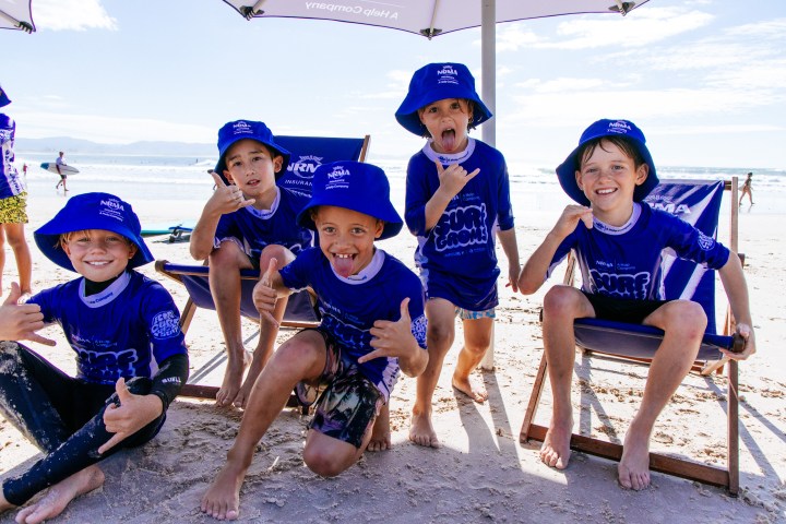 Five children in blue hats and shirts posing on a sunny beach with surfboards in the background.