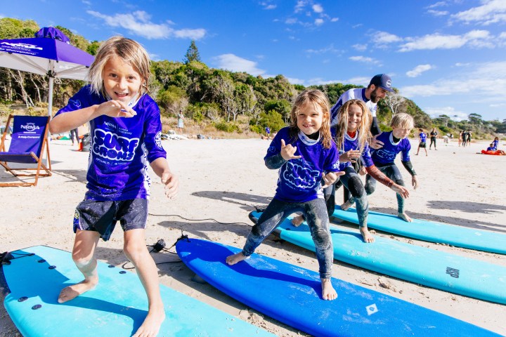 Children in purple shirts practice surfing on blue boards at a sunny beach.