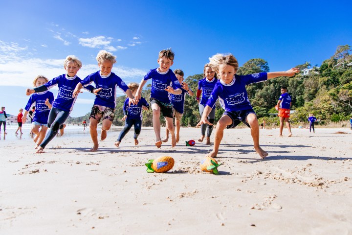 Children in blue shirts play with toy pineapples on a sunny beach.