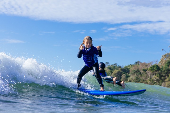 Young surfer on blue board riding a wave, smiling and making a shaka sign under a clear sky.