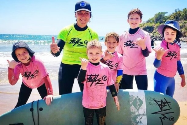 Group of kids and instructor in surf gear smiling and giving thumbs-up on a sunny beach.