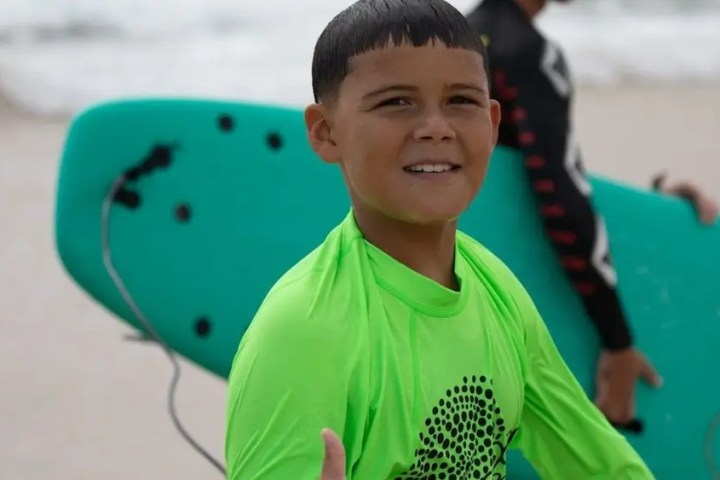 Young boy in green shirt smiles, holding a surfboard on the beach.